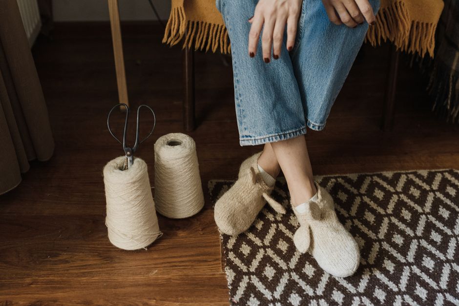 A person sitting on a wooden floor inside a residential room, wearing cozy knitted slippers and blue jeans with a mustard-colored shawl draped over their lap. Next to them are two spools of light-colo