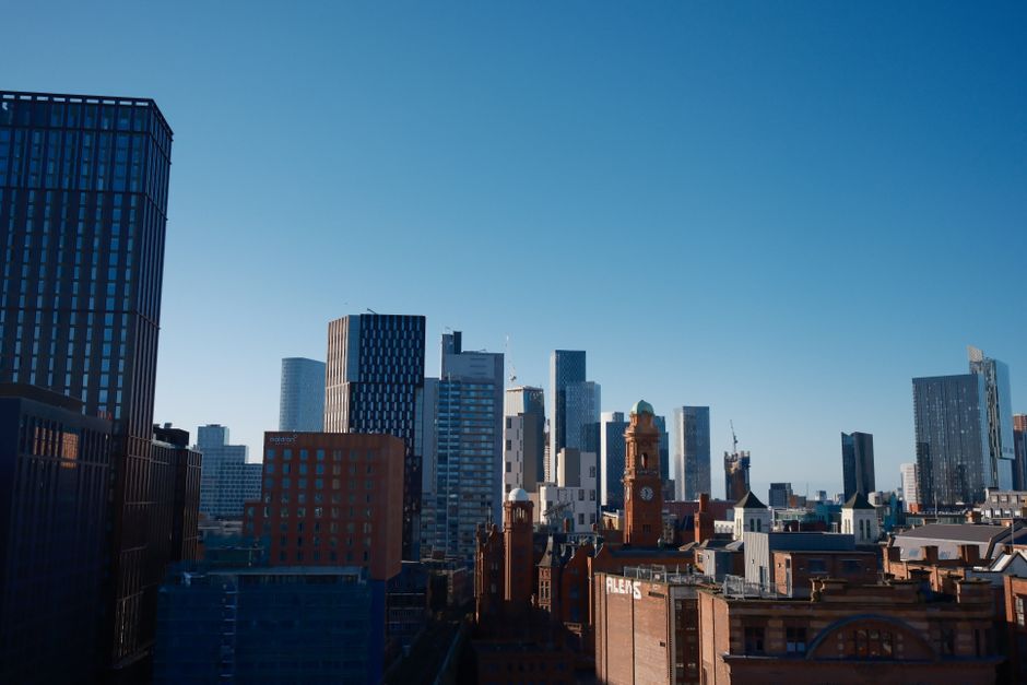 A city skyline featuring modern high-rise office buildings and historic brick structures in London, with a clear blue sky above. The scene includes glass-clad towers reflecting sunlight and traditiona