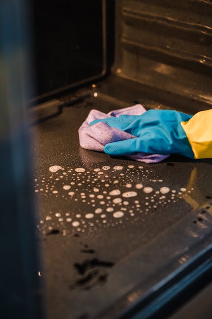 A close-up view of a white stove with a black metal grate and a beige tiled wall in the background. A person's hand wearing a yellow and blue rubber glove is using a purple cleaning cloth to wipe the 